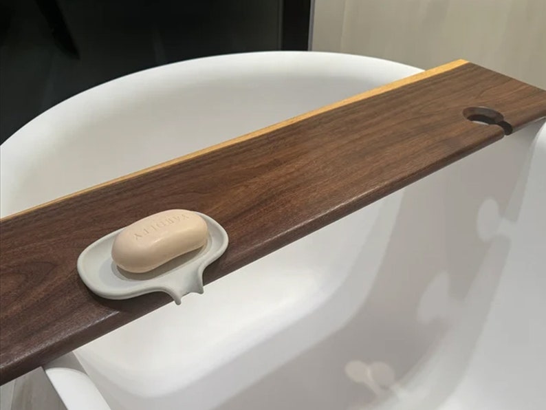 May include: A wooden bath tray with a soap dish and a hole for a glass. The tray is made of dark wood and has a light wood accent. The soap dish is white and has a soap bar inside.