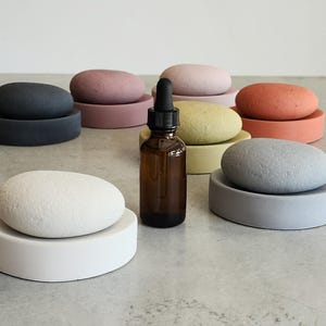 May include: A collection of colorful, oval-shaped soap dishes with matching stone-like soaps. The set includes dishes and soaps in various colors: white, black, pink, yellow, orange, and gray. A brown glass bottle with a dropper is in the center.