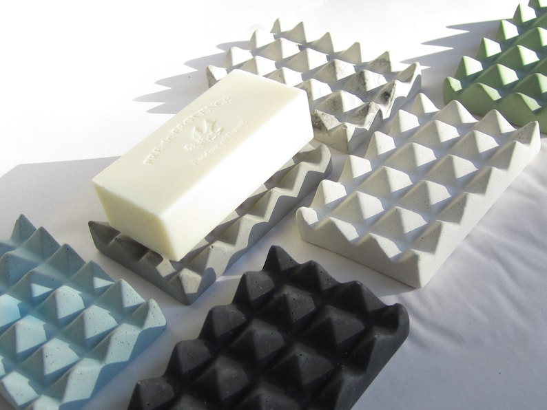 May include: Five concrete soap dishes in various colors, including gray, black, blue, and green. The dishes have a textured, pyramid-shaped design. A white bar of soap is resting on one of the dishes.