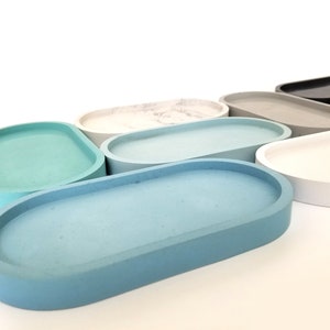 May include: A set of oval-shaped concrete trays in various colours, including light blue, teal, grey, white, and beige. The trays are arranged in a row, with the largest tray at the front and the smallest tray at the back.