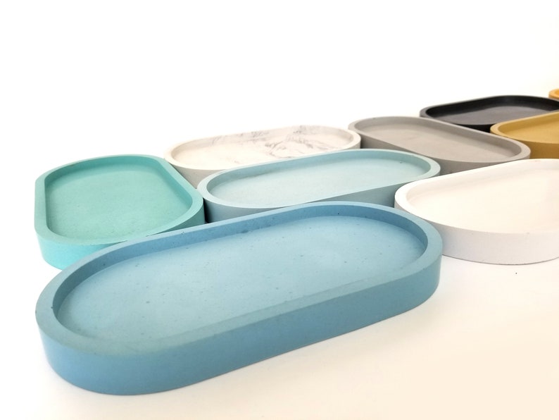 May include: A set of oval-shaped concrete trays in various colours, including light blue, teal, grey, white, and beige. The trays are arranged in a row, with the largest tray at the front and the smallest tray at the back.
