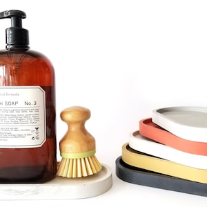 May include: A brown glass bottle of dish soap with a white label that reads "Botanical formula Dish Soap No. 3". The bottle is sitting on a grey concrete oval dish. Next to the bottle is a wooden brush with green bristles. There are five stacked oval concrete dishes in various colours, including grey, white, red, yellow, and black.