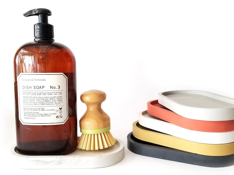 May include: A brown glass bottle of dish soap with a white label that reads "Botanical formula Dish Soap No. 3". The bottle is sitting on a grey concrete oval dish. Next to the bottle is a wooden brush with green bristles. There are five stacked oval concrete dishes in various colours, including grey, white, red, yellow, and black.