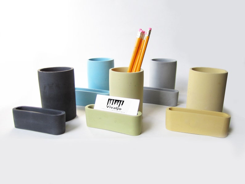 Op de afbeelding: Set van zes betonnen bureau-organizers in verschillende kleuren, waaronder grijs, lichtblauw, groen en geel. Elke organizer heeft een cilindrische vorm en een bijpassende rechthoekige kaarthouder. De set bevat een potloodhouder met een visitekaartje met de tekst "Vivajio".