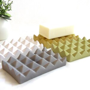 May include: Four ceramic soap dishes in white, gray, yellow, and green. Each dish has a textured surface with a raised diamond pattern. A white bar of soap is sitting on the yellow dish.