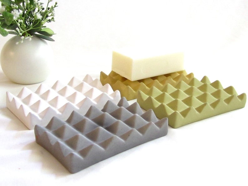 May include: Four ceramic soap dishes in white, gray, yellow, and green. Each dish has a textured surface with a raised diamond pattern. A white bar of soap is sitting on the yellow dish.