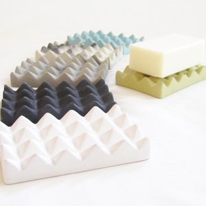 May include: A set of six soap dishes in various colors, including white, gray, black, and green. The dishes are made of a textured material and have a geometric design. A bar of white soap is sitting on top of one of the green dishes.