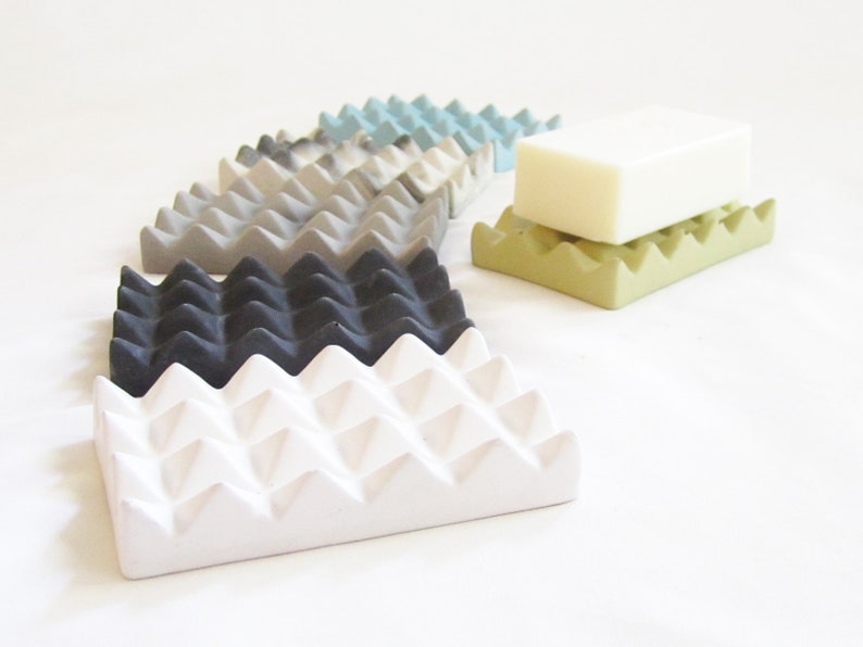 May include: A set of six soap dishes in various colors, including white, gray, black, and green. The dishes are made of a textured material and have a geometric design. A bar of white soap is sitting on top of one of the green dishes.