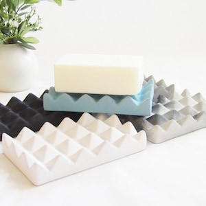 May include: Four soap dishes made of concrete in different colors: black, white, light blue, and gray with a marbled pattern. The dishes have a unique design with triangular ridges.
