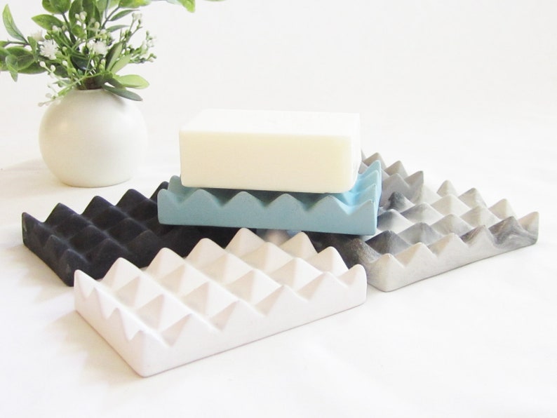 May include: Four soap dishes made of concrete in different colors: black, white, light blue, and gray with a marbled pattern. The dishes have a unique design with triangular ridges.