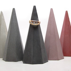 May include: Five concrete ring holders in different colors: gray, light gray, pink, and red. The ring holders are shaped like pyramids with a flat top.