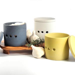 May include: Three ceramic garlic keepers with lids. The keepers are in grey, white, and yellow. The lids are off of the grey and yellow keepers. There are garlic cloves and rosemary sprigs in front of the keepers.