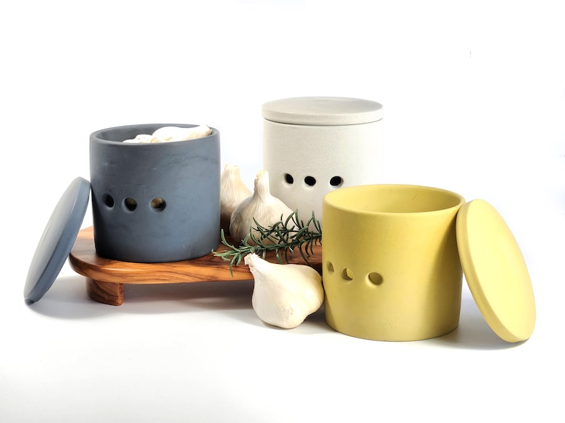 May include: Three ceramic garlic keepers with lids. The keepers are in grey, white, and yellow. The lids are off of the grey and yellow keepers. There are garlic cloves and rosemary sprigs in front of the keepers.