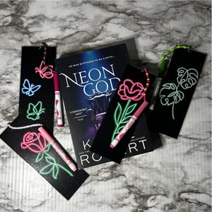 May include: A book titled "Neon God" with black bookmarks featuring neon-style designs of a rose, butterflies, and a plant. Pink and green markers are placed near the bookmarks. The book cover has the text "He was supposed to be a myth..."