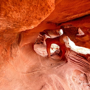 May include: A close-up view of a red sandstone cave with a natural archway. The cave is lit by sunlight streaming through the opening.