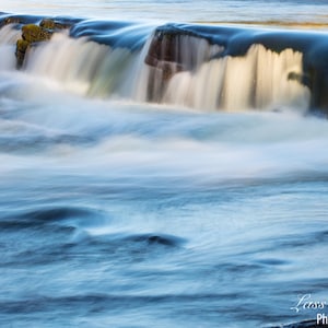 May include: A close-up of a waterfall with white water flowing over rocks. The water is blurred due to a long exposure, creating a silky smooth effect. The rocks are covered in moss and are a dark green color.