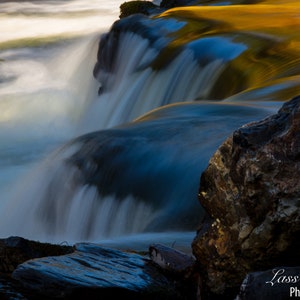 May include: A close-up of a waterfall with a blue and white water flow over rocks. The water is blurred due to long exposure, creating a silky smooth effect. The rocks are dark brown and gray.