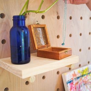 May include: A wooden shelf with a blue glass bottle, a small wooden box with a glass lid, and a blue and white eye charm necklace hanging from the shelf.