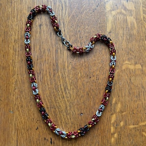 May include: A handmade necklace composed of small, interlocking metal rings. The necklace features a color scheme of red, black, gold, and silver. The chain is laid out on a wooden surface.