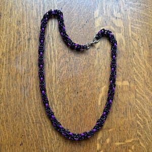 May include: A handmade necklace featuring a Byzantine chainmaille design. The chain is composed of black and purple anodized aluminum rings, creating a striking contrast. The necklace is secured with a silver-colored clasp and rests on a wooden surface.