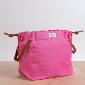 May include: A bright pink tote bag with brown leather handles and drawstring closure. The bag has a rectangular shape with a small white label on the front. The bag is sitting on a light brown wooden surface.