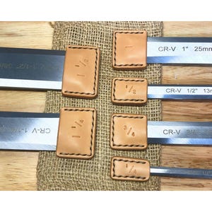 May include: A set of six metal chisels with tan leather blade guards. Each guard is stamped with a fraction size, including 1 1/2, 1 1/4, 1, 1/2, 3/4, and 1/4. The steel blades are marked CR-V with corresponding imperial and metric measurements.
