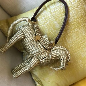 Pu&ograve; includere: Una borsa a mano intrecciata color oro con un manico in pelle marrone scuro. La borsa ha una forma unica e una chiusura color oro. La borsa &egrave; appoggiata su un cuscino giallo.
