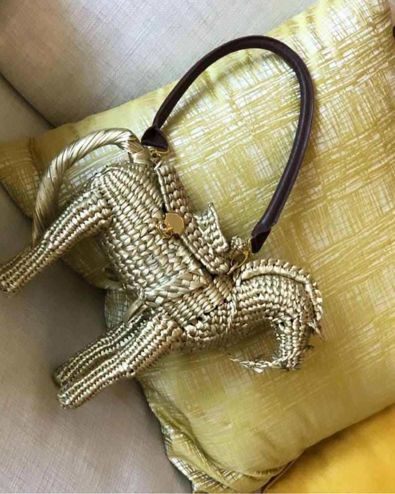 Pu&ograve; includere: Una borsa a mano intrecciata color oro con un manico in pelle marrone scuro. La borsa ha una forma unica e una chiusura color oro. La borsa &egrave; appoggiata su un cuscino giallo.