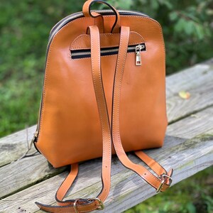 May include: A tan leather backpack with adjustable straps and a zipper closure. The backpack is sitting on a wooden surface.