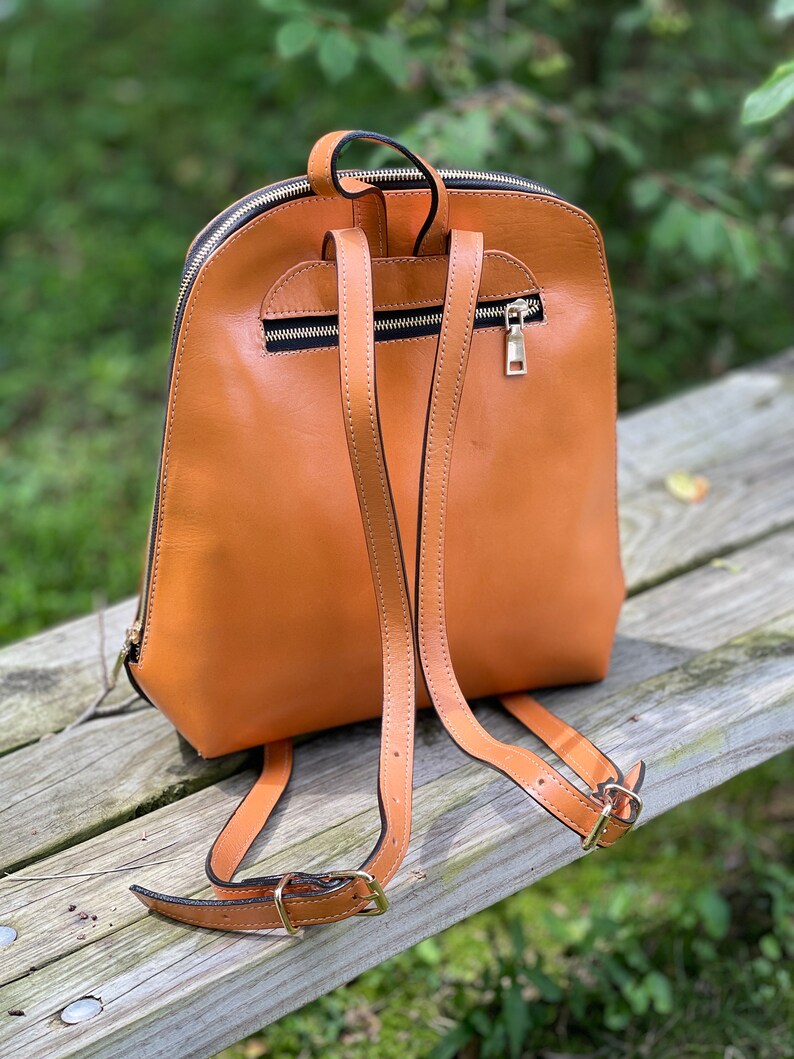 May include: A tan leather backpack with adjustable straps and a zipper closure. The backpack is sitting on a wooden surface.