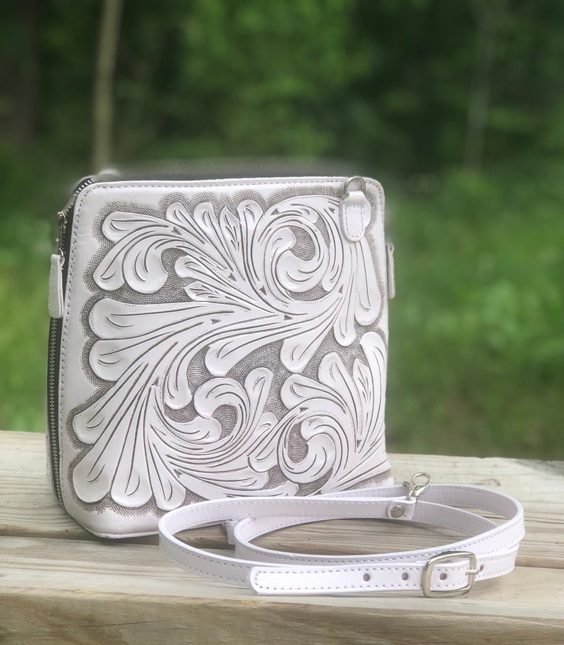 Peut inclure: Sac &agrave; bandouli&egrave;re en cuir blanc avec un motif floral complexe en relief sur le devant. Le sac a une bandouli&egrave;re r&eacute;glable blanche avec une boucle argent&eacute;e.