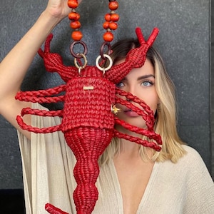 Peut inclure: Un sac à main rouge vif en forme de homard tressé. Le sac est doté d'une anse ornée de perles en bois orange et d'une lanière en cuir marron foncé. Des anneaux dorés et un petit fermoir doré ajoutent des détails. Le design comprend des pinces, des pattes et une queue.