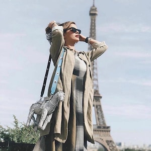 Pu&ograve; includere: Una persona con occhiali da sole, un lungo cappotto verde oliva e un vestito a quadri &egrave; in piedi davanti alla Torre Eiffel. Una borsa a forma di cavallo argentato con una tracolla azzurra &egrave; appesa alla sua spalla.