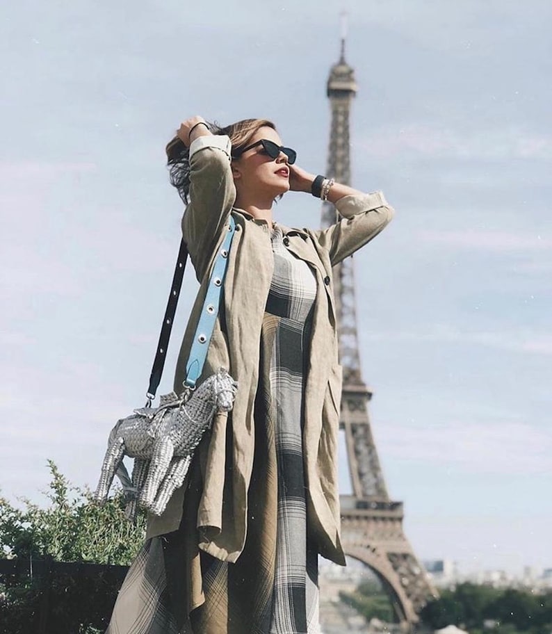 Pu&ograve; includere: Una persona con occhiali da sole, un lungo cappotto verde oliva e un vestito a quadri &egrave; in piedi davanti alla Torre Eiffel. Una borsa a forma di cavallo argentato con una tracolla azzurra &egrave; appesa alla sua spalla.