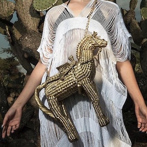Pu&ograve; includere: Una borsa a forma di cavallo intrecciata color oro con una tracolla a catena. La borsa ha dettagli di sella e briglia. La persona indossa un copricostume bianco con frange. Lo sfondo presenta un cactus.