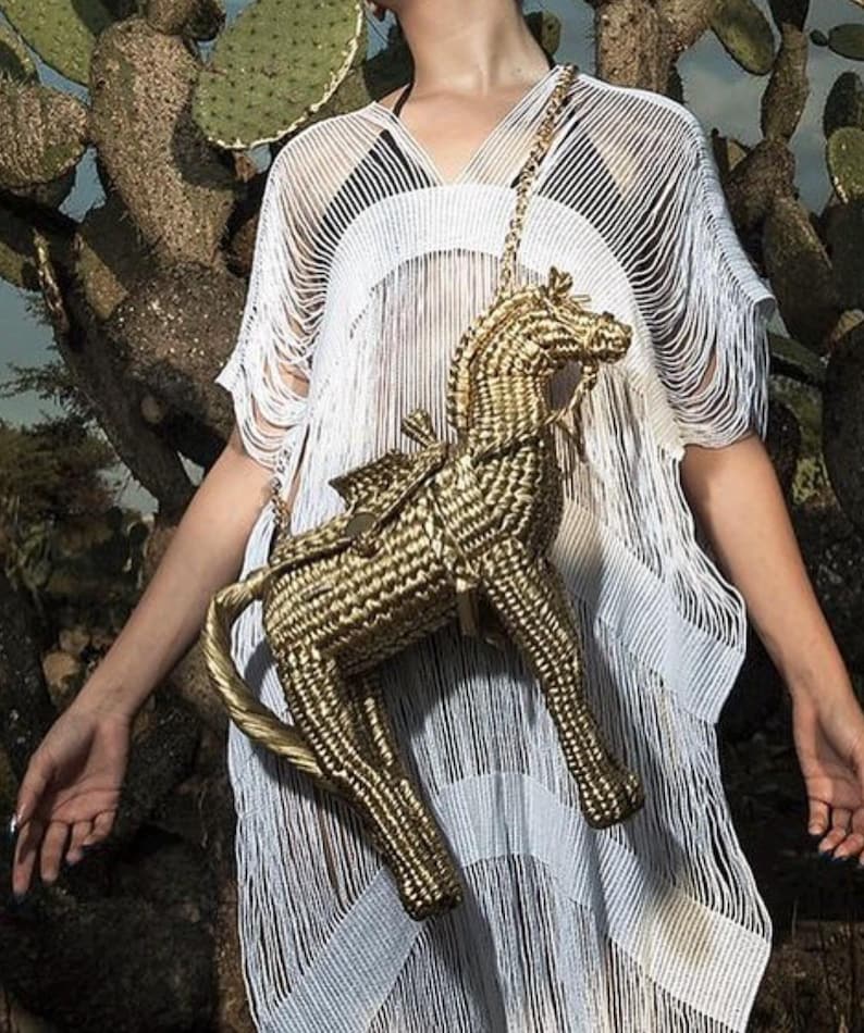 Pu&ograve; includere: Una borsa a forma di cavallo intrecciata color oro con una tracolla a catena. La borsa ha dettagli di sella e briglia. La persona indossa un copricostume bianco con frange. Lo sfondo presenta un cactus.