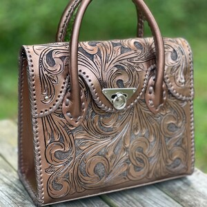 Puede incluir: Un bolso de mano de cuero marr&oacute;n con intrincados dise&ntilde;os florales tallados. El bolso tiene un asa superior y un cierre plateado.