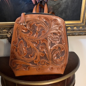 May include: A brown leather backpack with intricate floral tooling. The backpack has a zipper closure and a small tassel.
