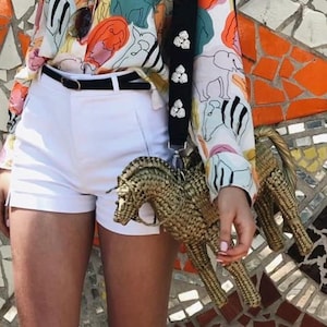 Pu&ograve; includere: Una borsa a forma di cavallo intrecciata con un cinturino nero adornato con abbellimenti bianchi. La persona indossa pantaloncini bianchi, una cintura nera e una camicetta fantasia colorata con disegni di animali e astratti. Sono visibili occhiali da sole.