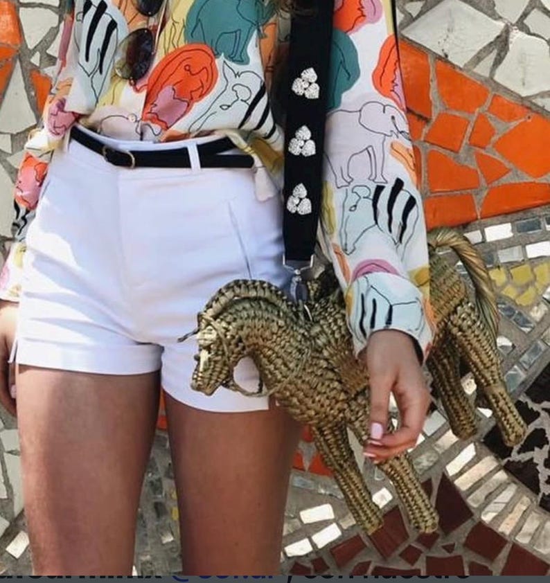 Pu&ograve; includere: Una borsa a forma di cavallo intrecciata con un cinturino nero adornato con abbellimenti bianchi. La persona indossa pantaloncini bianchi, una cintura nera e una camicetta fantasia colorata con disegni di animali e astratti. Sono visibili occhiali da sole.