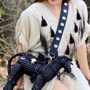 Pu&ograve; includere: Una borsa a tracolla nera a forma di cavallo intrecciato con chiusura argentata e cinturino nero ornato di borchie argentate. La borsa &egrave; indossata sopra un vestito color crema con dettagli a nappe nere.
