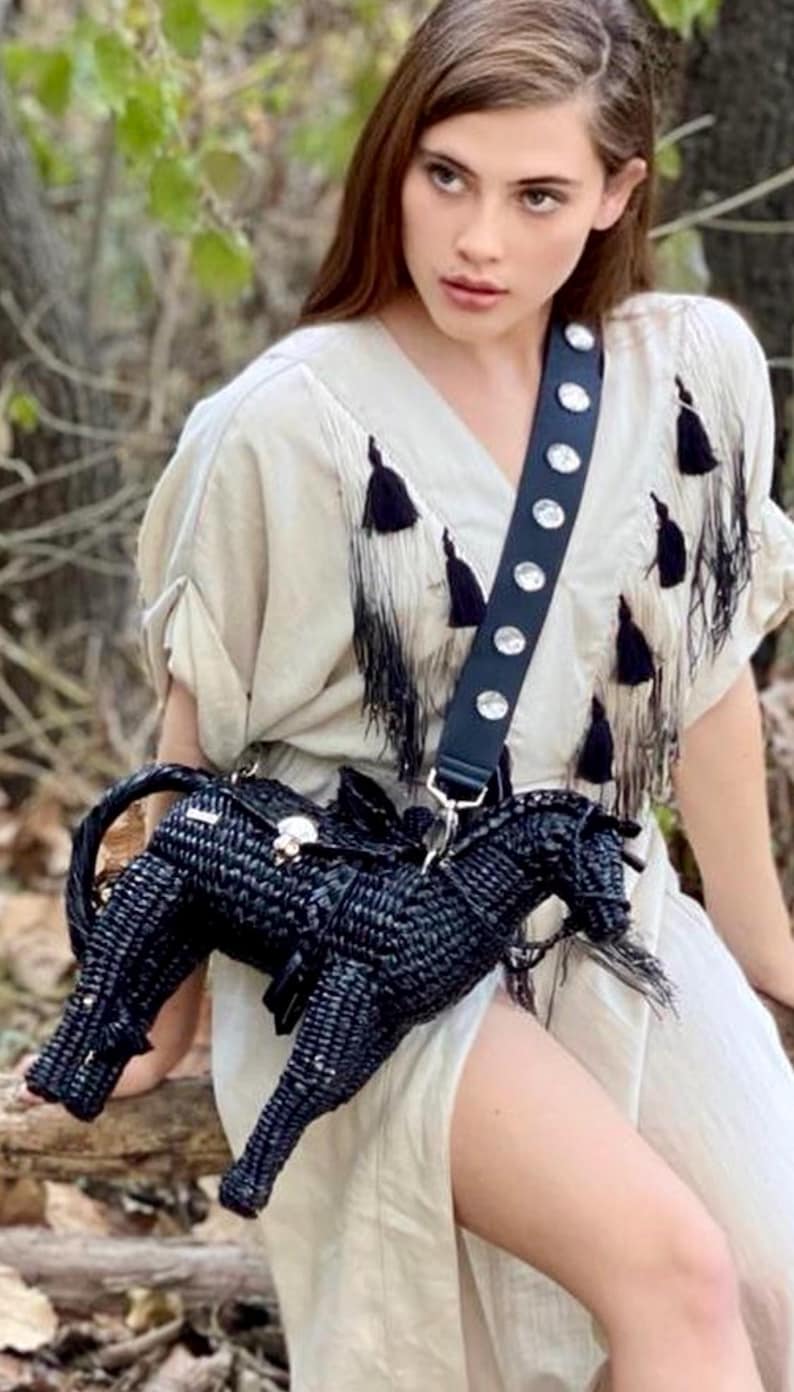 Pu&ograve; includere: Una borsa a tracolla nera a forma di cavallo intrecciato con chiusura argentata e cinturino nero ornato di borchie argentate. La borsa &egrave; indossata sopra un vestito color crema con dettagli a nappe nere.