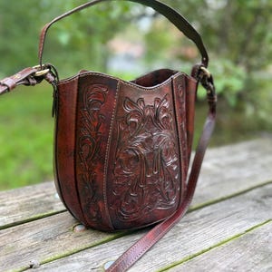 May include: A dark brown leather bucket bag featuring intricate floral tooling and embossed scrollwork patterns. The bag has a structured, rounded base, a single top handle, and a long adjustable shoulder strap attached with brass-toned metal hardware.