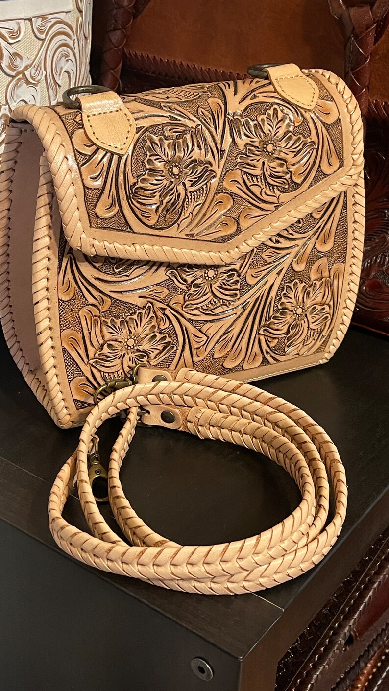 Pode incluir: Uma bolsa de couro marrom claro com um design floral trabalhado e uma al&ccedil;a tran&ccedil;ada. A bolsa tem um fecho de aba e um pequeno bolso na frente.