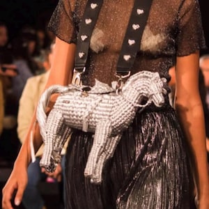 Pu&ograve; includere: Una borsa a forma di cavallo argentata e bianca con una tracolla nera ornata di borchie bianche a forma di cuore. La borsa &egrave; indossata sopra una gonna nera plissettata e un top trasparente e glitterato. Lo stile generale &egrave; alla moda.