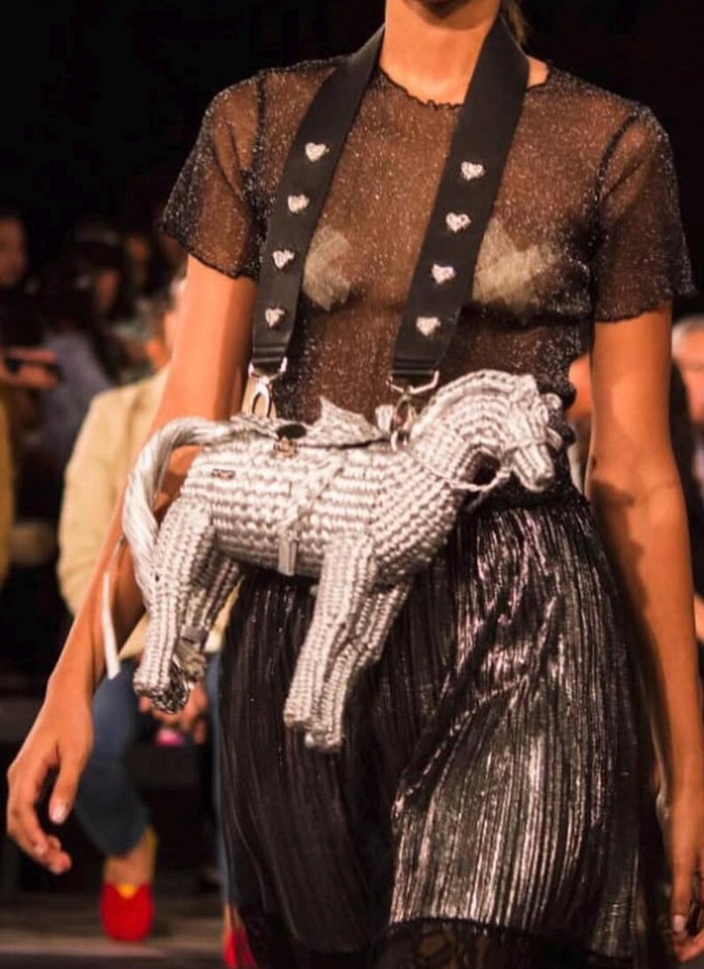 Pu&ograve; includere: Una borsa a forma di cavallo argentata e bianca con una tracolla nera ornata di borchie bianche a forma di cuore. La borsa &egrave; indossata sopra una gonna nera plissettata e un top trasparente e glitterato. Lo stile generale &egrave; alla moda.