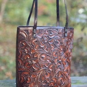 May include: A brown leather tote bag with a floral embossed design. The bag has two straps and a top zipper closure.