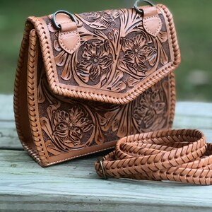 Pode incluir: Uma bolsa de couro marrom transversal com detalhes florais intrincados. A bolsa tem um fecho de aba e uma al&ccedil;a de couro tran&ccedil;ada.
