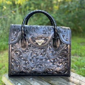 Puede incluir: Un bolso de mano de cuero negro con un intrincado dise&ntilde;o floral y un asa trenzada. El bolso tiene un cierre plateado y est&aacute; sobre una superficie de madera.