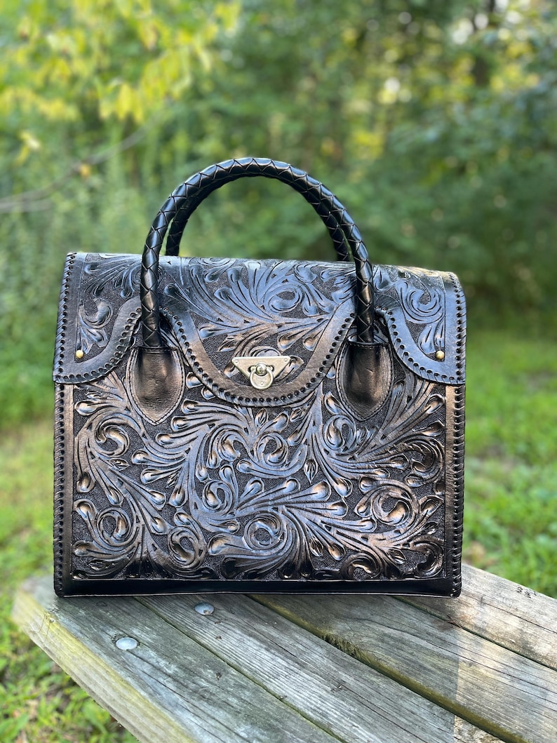 Puede incluir: Un bolso de mano de cuero negro con un intrincado dise&ntilde;o floral y un asa trenzada. El bolso tiene un cierre plateado y est&aacute; sobre una superficie de madera.